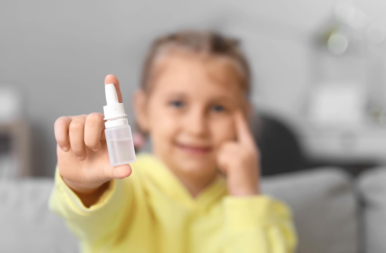 A young girl points to her eye with her left hand, holding an unmarked bottle of eye drops up in front of the camera.