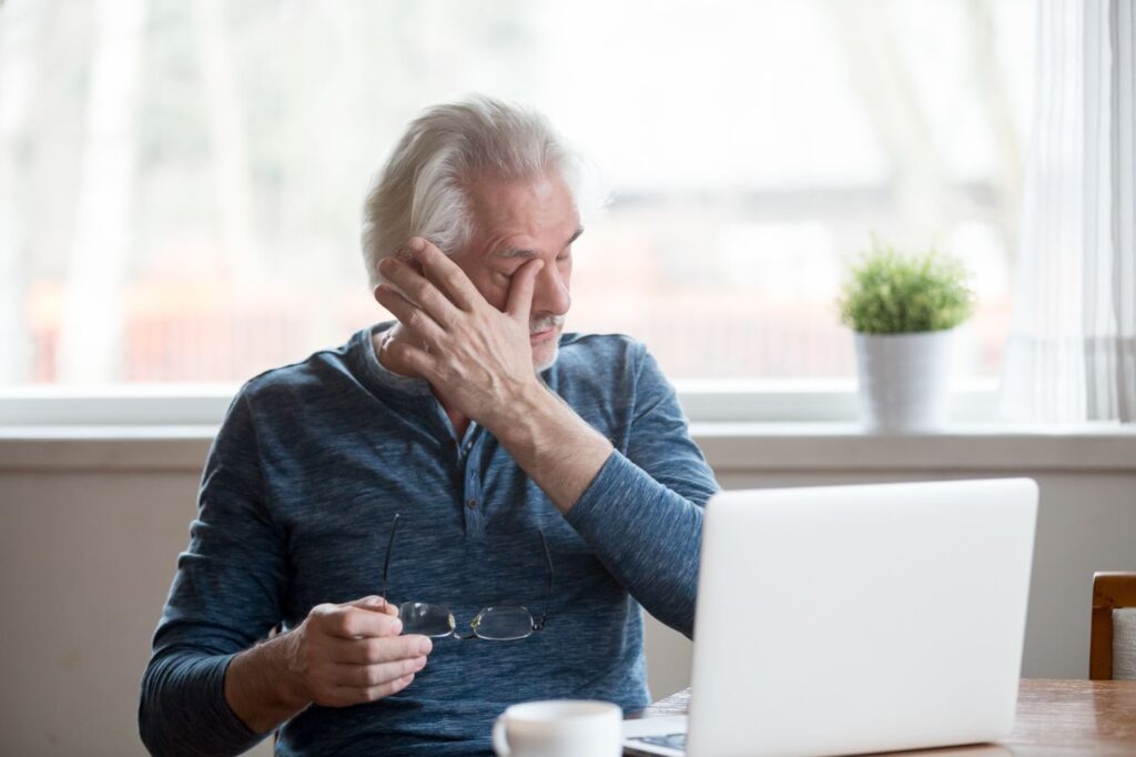 An older adult sitting in front of a laptop taking their glasses off and rubbing their eyes.