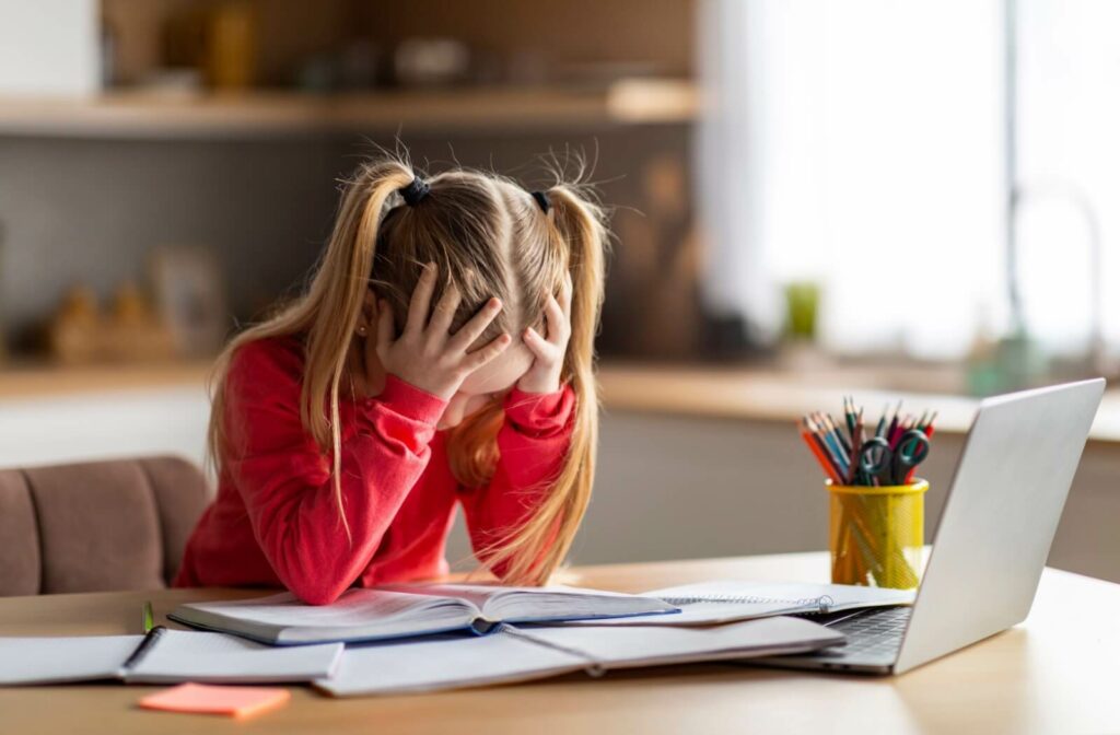 A young child with myopia sits at a desk. The put their head in their hands as they struggle to read a book on their desk.