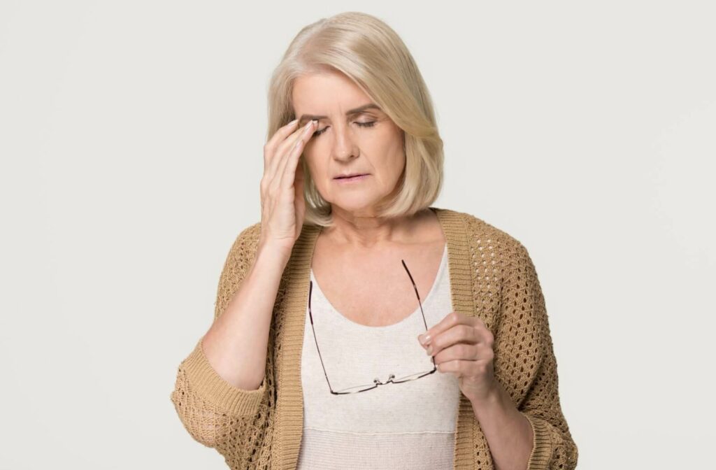 An older adult putting their right hand to their right eye and temple from a headache and holding eyeglasses in their left hand.