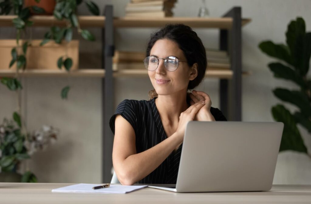 A smiling adult sitting in from of a laptop and looking away front he screen to rest their eyes.